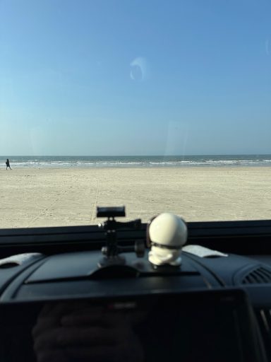 Blick vom Fahrzeuginneren auf den Strand und das Meer unter klarem Himmel.