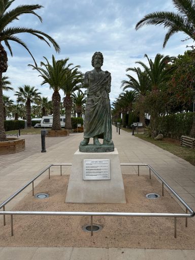 Eine bronzene Statue einer stehenden Person vor Palmen entlang eines gepflasterten Wegs.