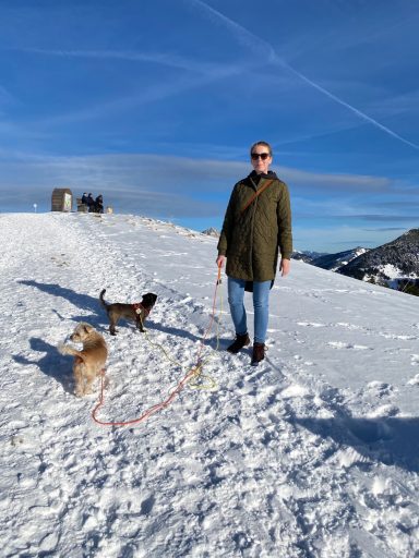 Mann in Winterkleidung steht auf Schnee mit zwei Hunden. Hintergrund zeigt Berge und blauen Himmel.