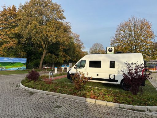 Weißer Wohnmobil in einem ruhigen Park mit Bäumen und einer Wandbild im Hintergrund.