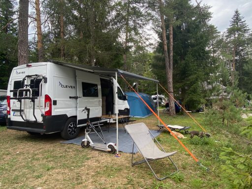 Wohnmobil auf einem Campingplatz, umgeben von Bäumen, mit Campingequipment und einem Rollstuhl.
