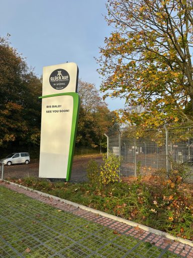 Schild mit dem Namen einer Einrichtung, umgeben von Bäumen und einem Parkplatz.