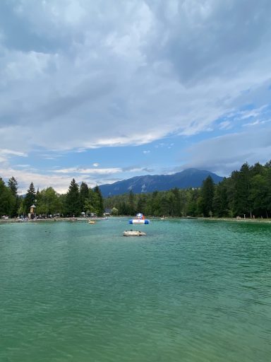 Blick auf einen klaren See umgeben von Bäumen und Bergen unter einem bewölkten Himmel.
